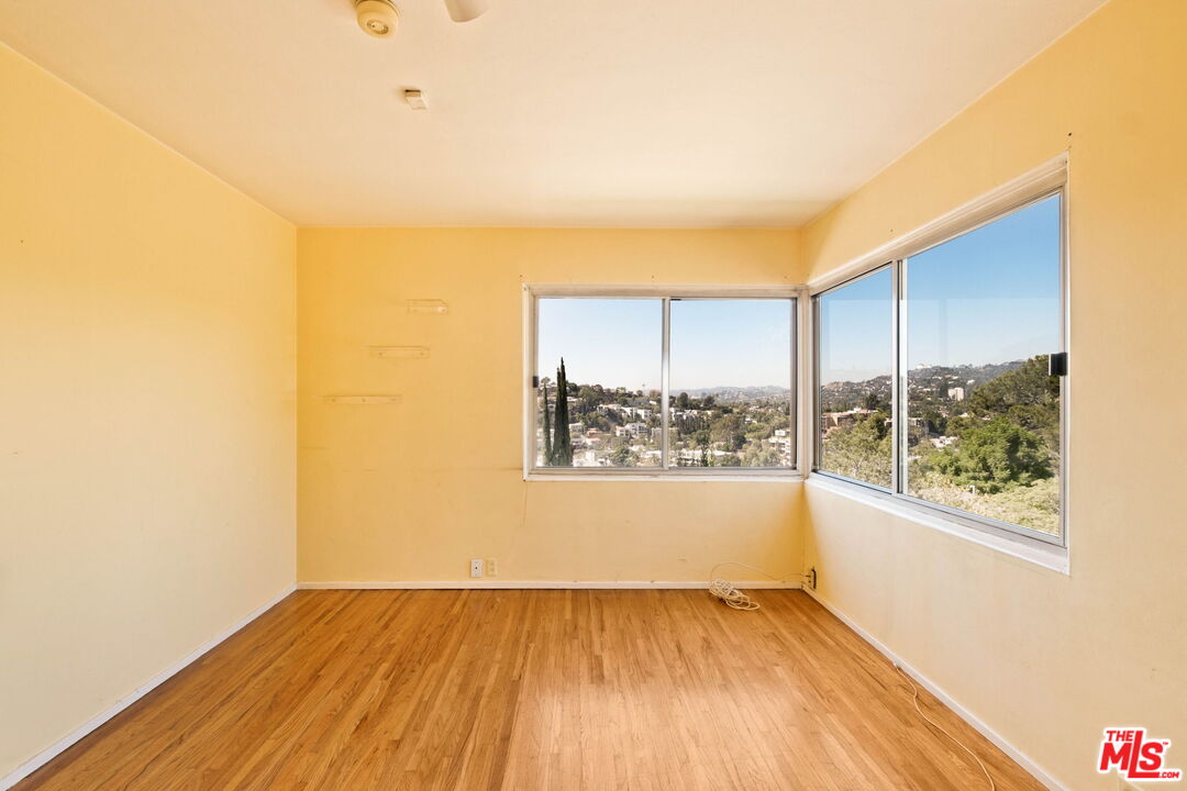 Vacant room with hardwood floors and corner windows overlooking a leafy hillside neighborhood.