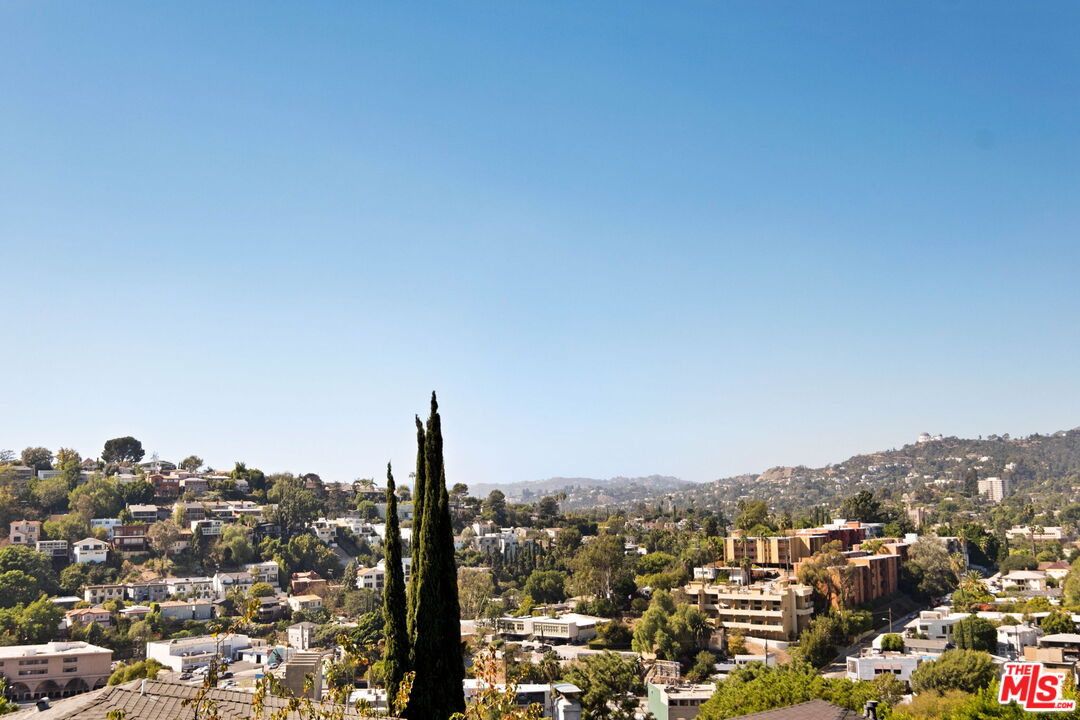 Panoramic view of hillside homes and distant hills, framed by two tall cypress trees.
