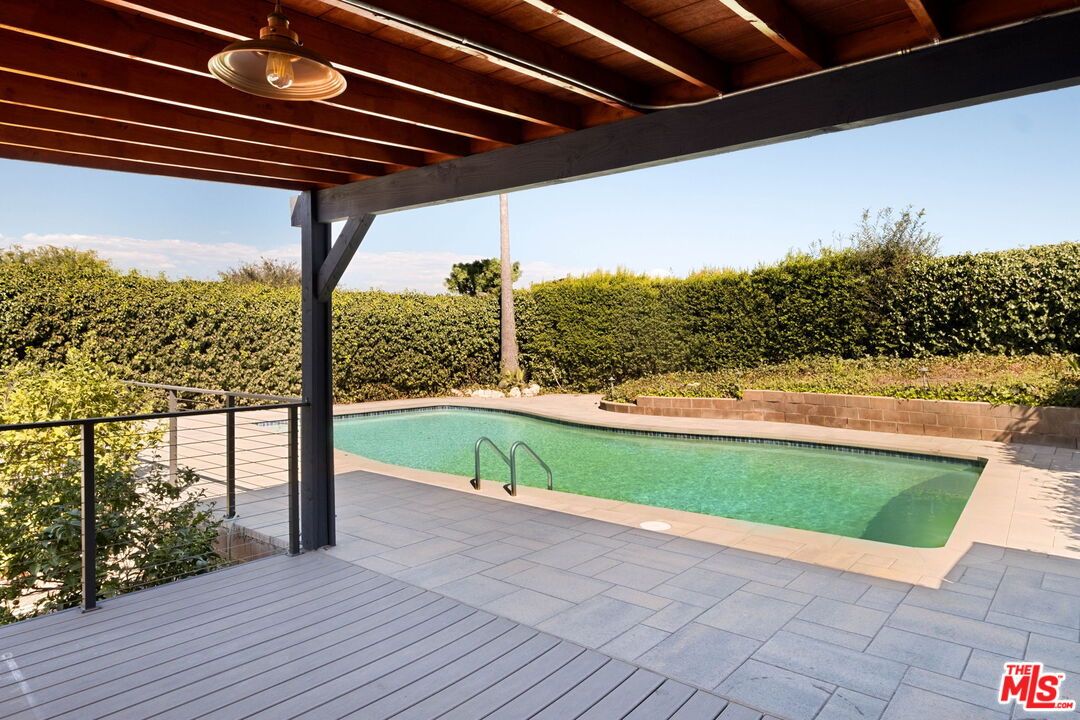 Backyard swimming pool beneath a wood-beamed pergola, surrounded by a paved patio and tall hedges.