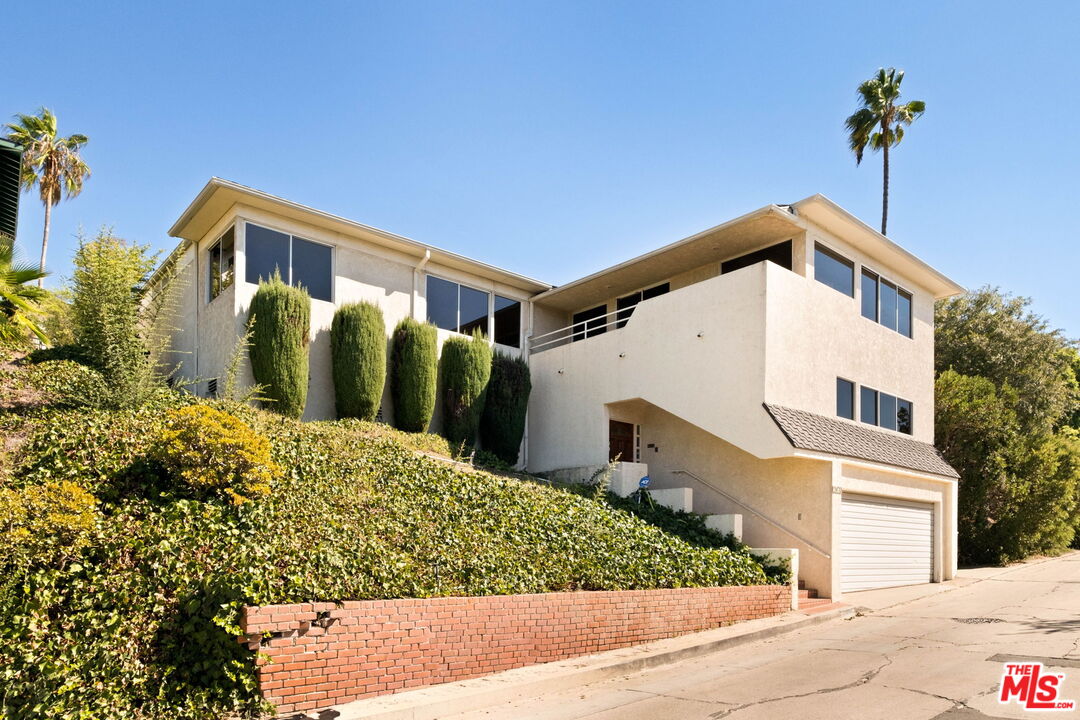 Modern cream stucco home with large windows, landscaped hillside, brick retaining wall, and attached garage.