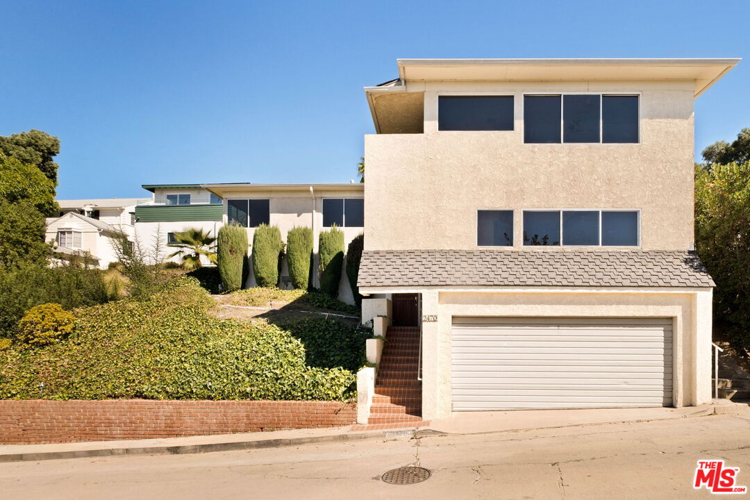 Multi-level stucco home with a wide garage and brick entry stairs beside a sloped landscaped yard.