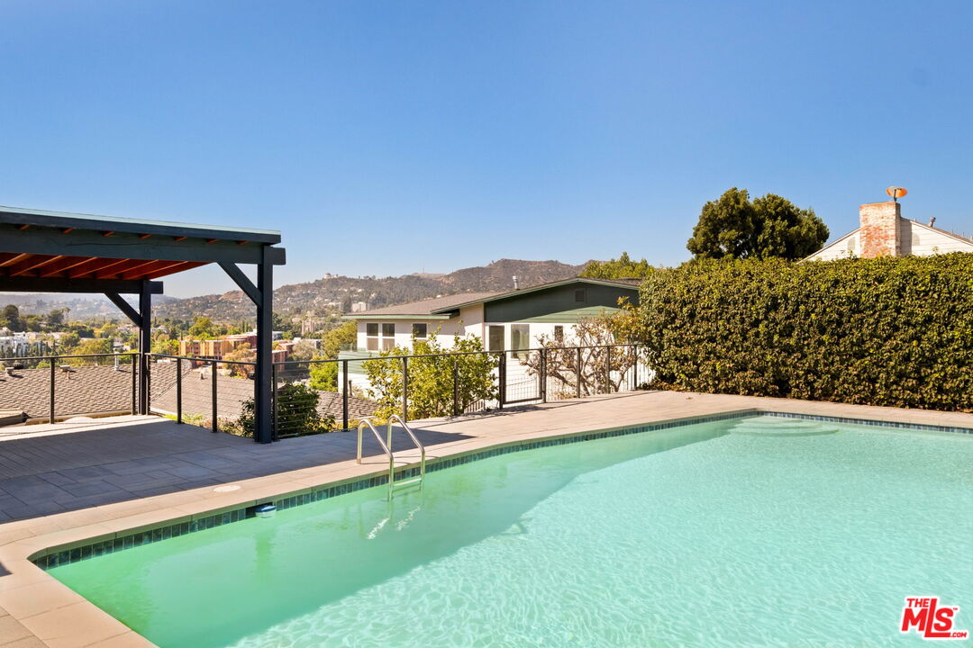 Swimming pool beside a covered patio, with a tall hedge and distant hills.