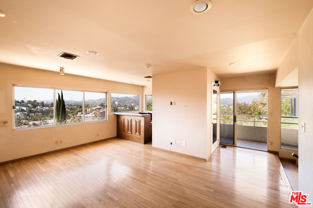 Empty living and dining area with wood floors, large hillside-view windows, and sliding doors to a balcony.