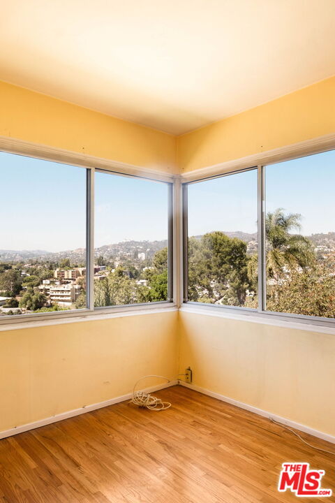 Empty room with hardwood floors and corner windows overlooking a leafy hillside neighborhood.