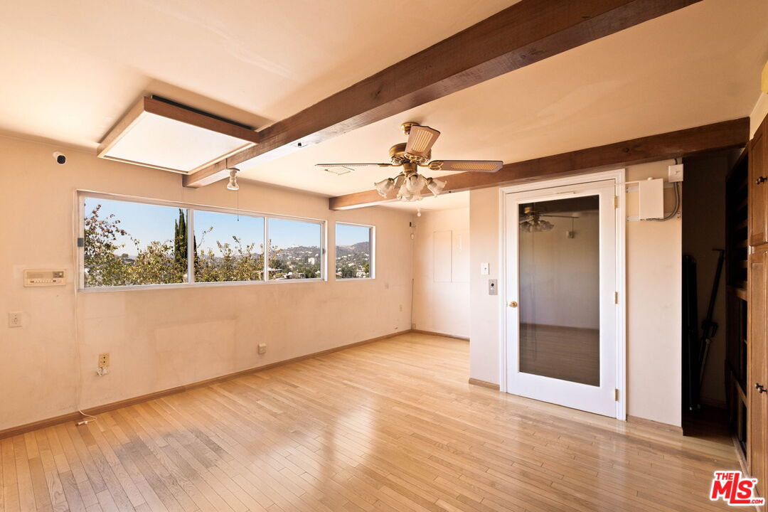 Empty room with wood floors, exposed ceiling beams, and windows overlooking hillside homes.