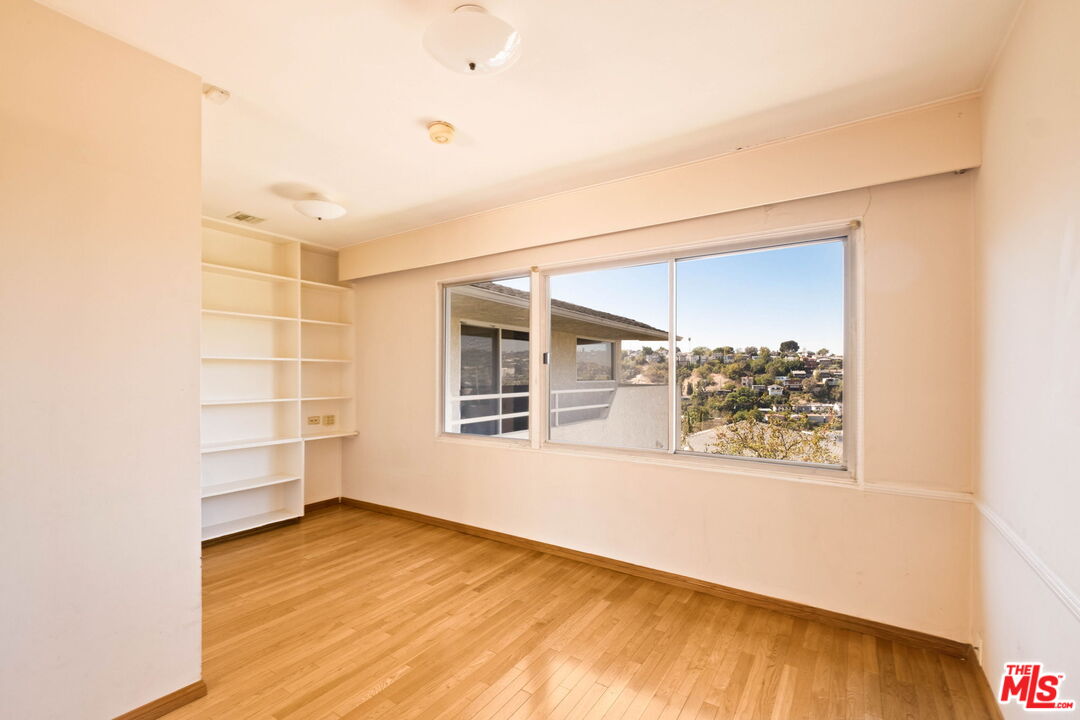 Empty room with hardwood floors, built-in shelving, and a large window overlooking a hillside neighborhood.