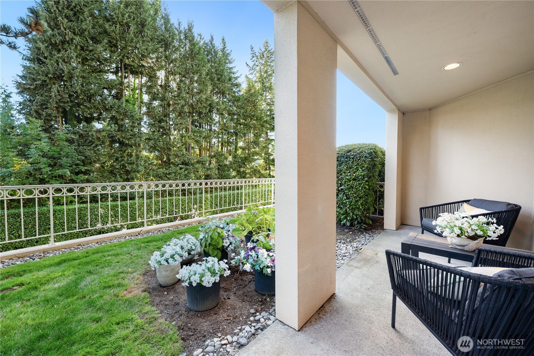 Covered patio with black outdoor seating beside a flower-filled garden and lawn.