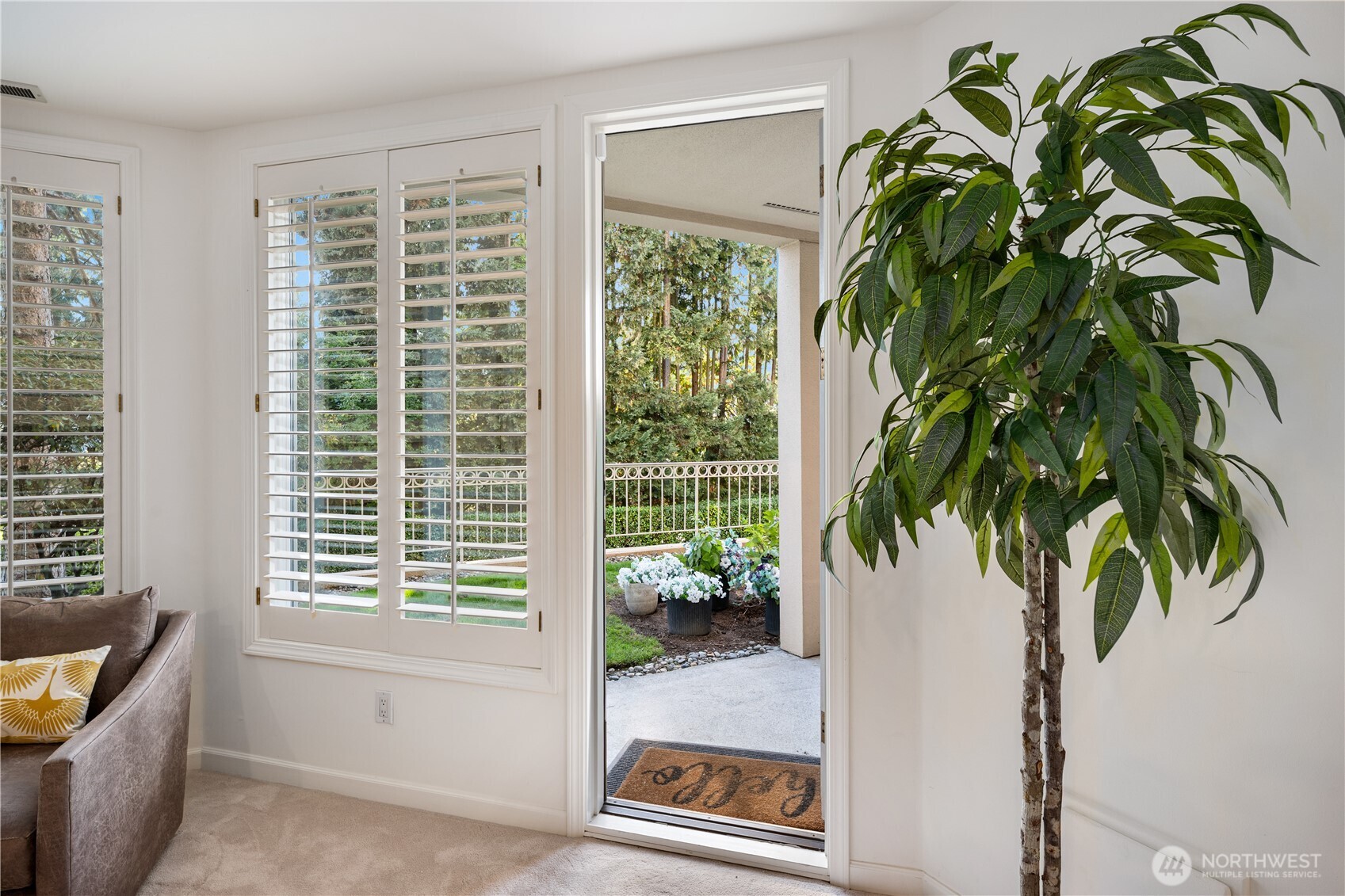 Living room with plantation shutters and an open patio doorway overlooking potted flowers.