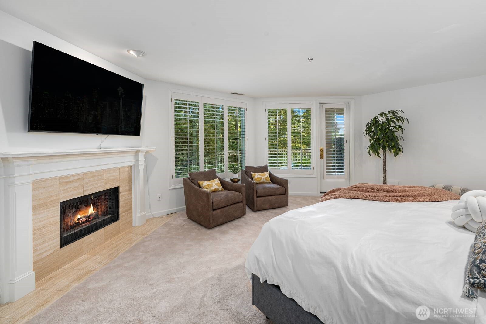 Bedroom with a lit tiled fireplace, two armchairs, and shuttered windows overlooking greenery.