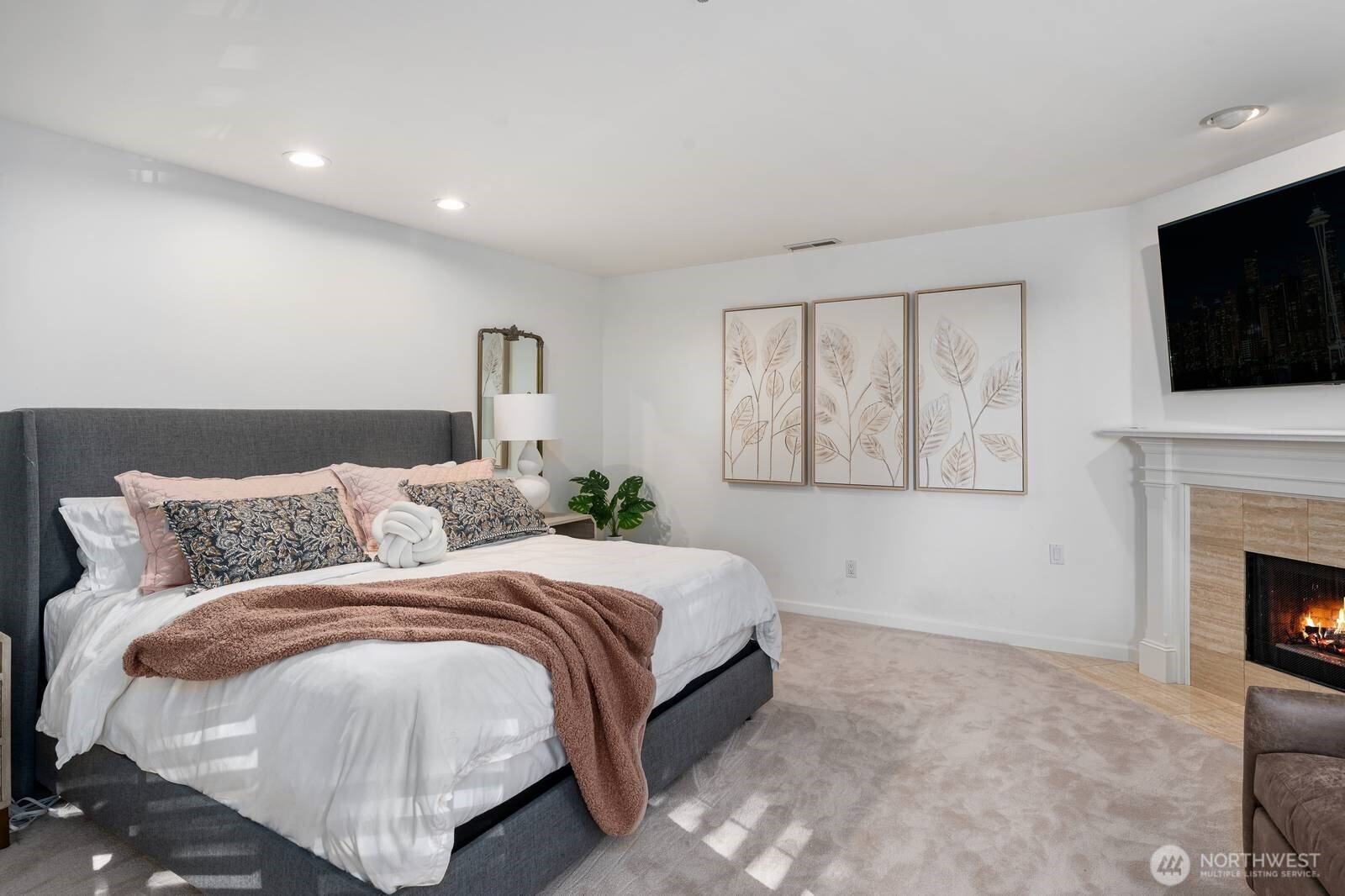 Staged bedroom with a gray upholstered bed, botanical prints, and a fireplace.