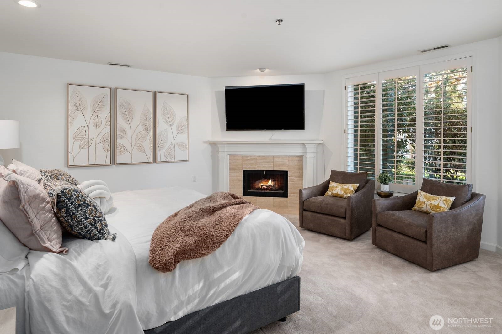 Staged bedroom with a fireplace, wall-mounted TV, two armchairs, and plantation-shuttered windows.