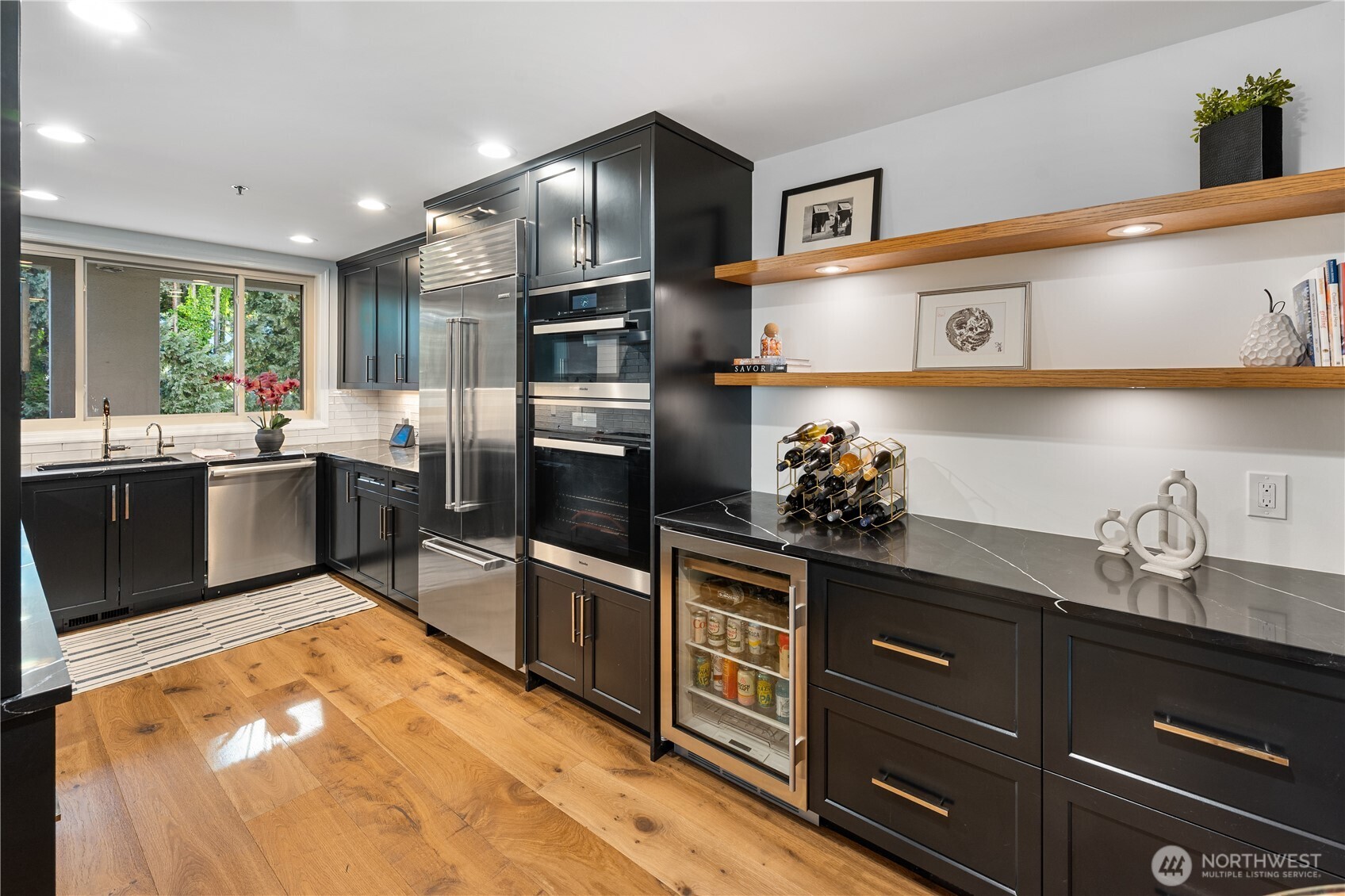 Modern kitchen with dark cabinets, stainless appliances, wood shelves, and a window overlooking greenery.