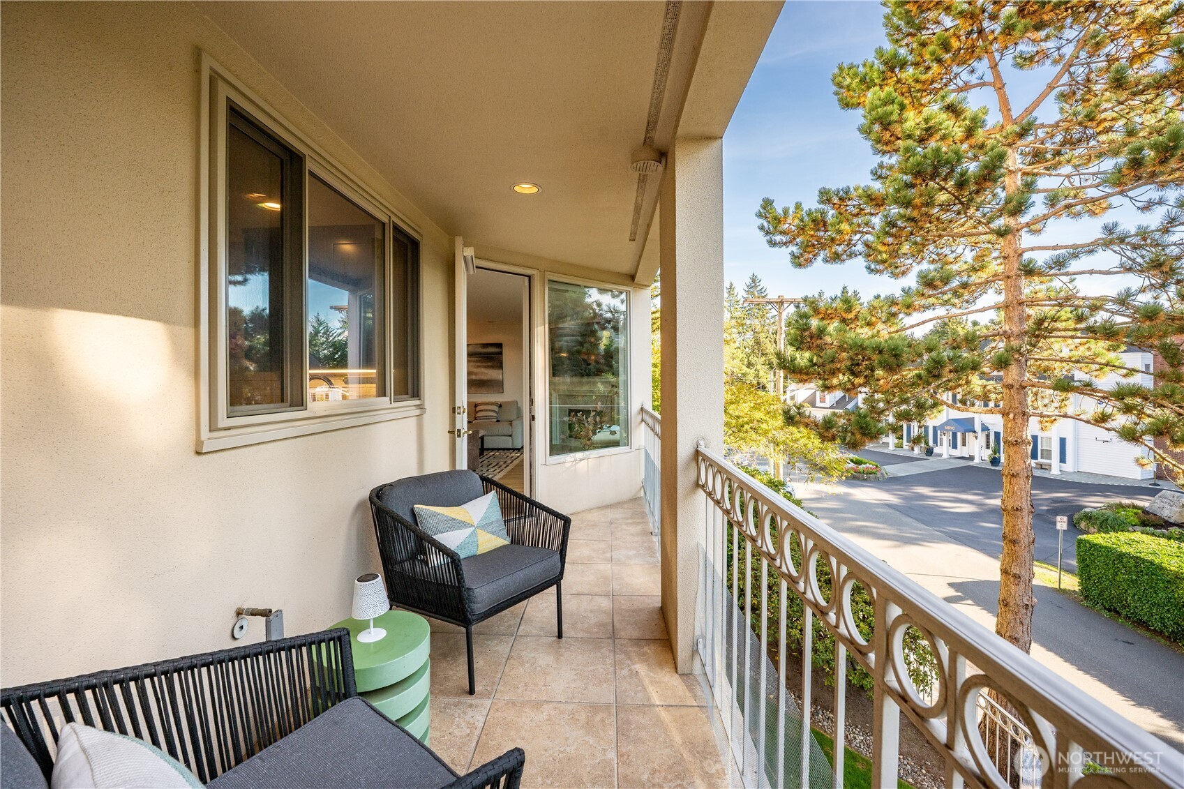 Furnished condo balcony with tiled flooring, woven chairs, and a view of a tree-lined street.