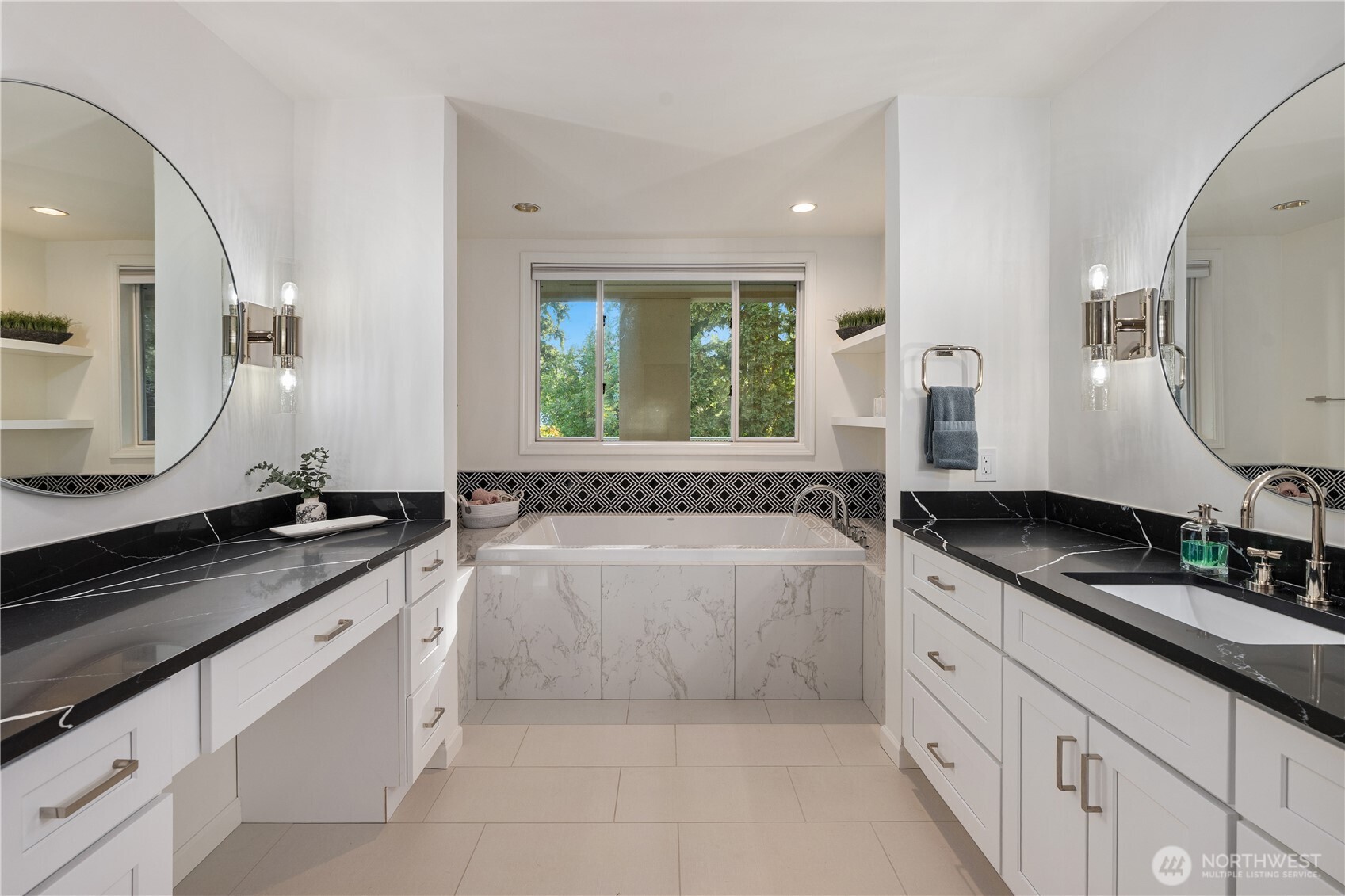 Bathroom with a built-in soaking tub, dual white vanities, black countertops, and large round mirrors.