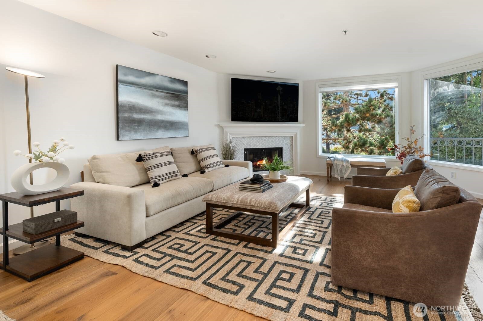 Furnished living room with a fireplace, large windows, and seating arranged around a patterned rug.