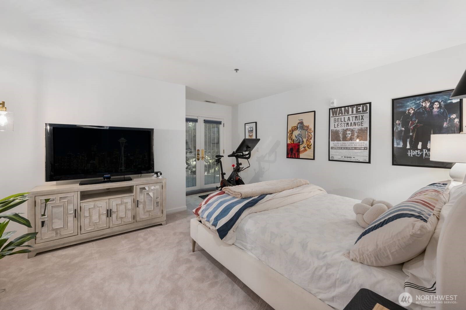 Furnished bedroom with striped bedding, framed posters, and an exercise bike beside glass doors.