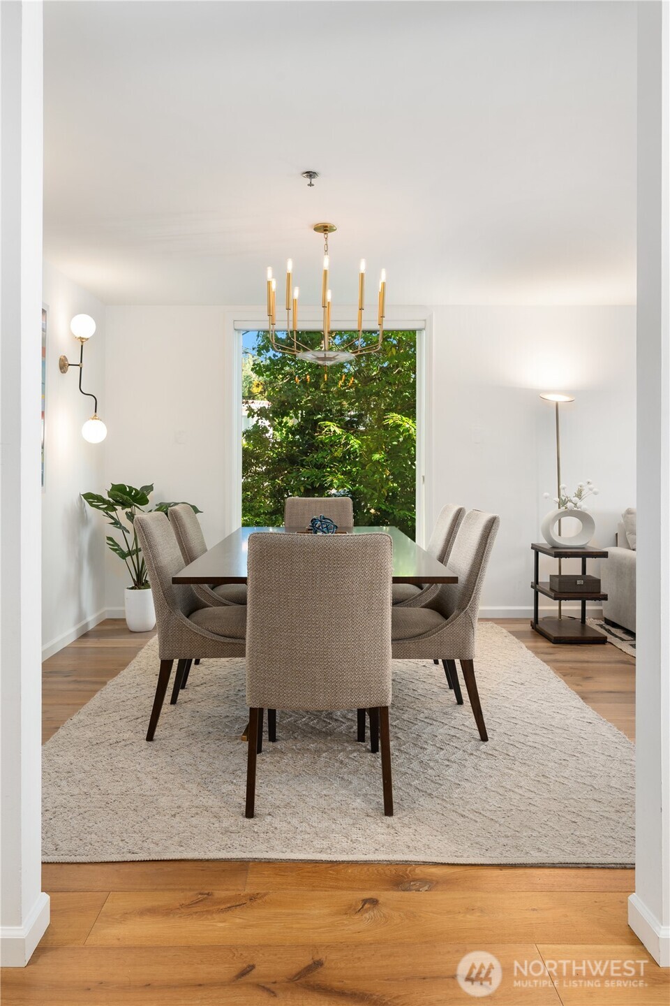 Dining room with upholstered chairs, a brass chandelier, and a large window overlooking leafy greenery.