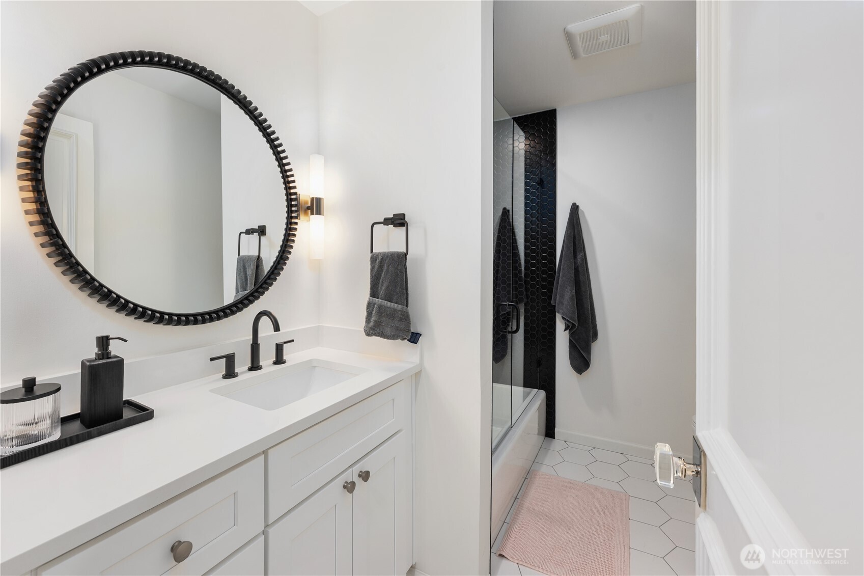 Bathroom with a white vanity, oversized round mirror, and glass shower with black hexagonal tile.