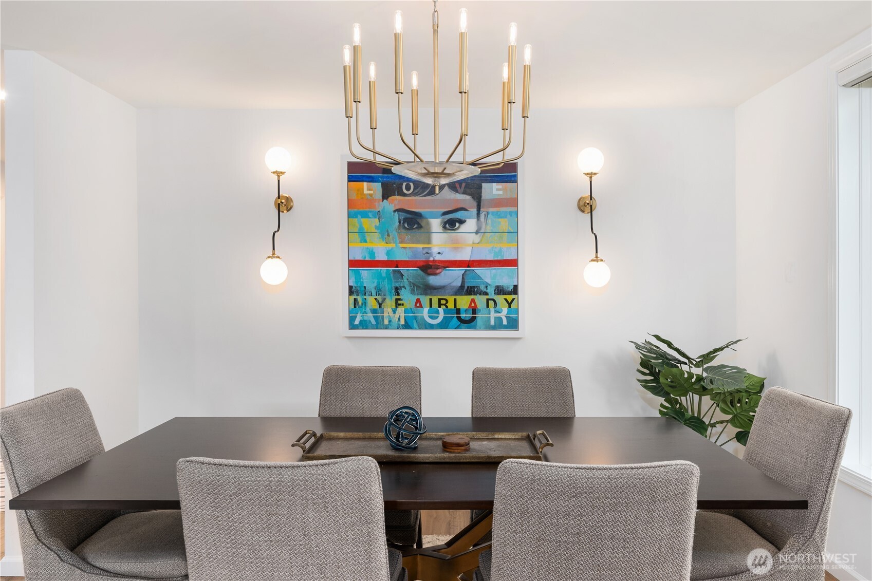 Dining room with a dark table, gray chairs, brass lighting, colorful portrait artwork, and a leafy plant.