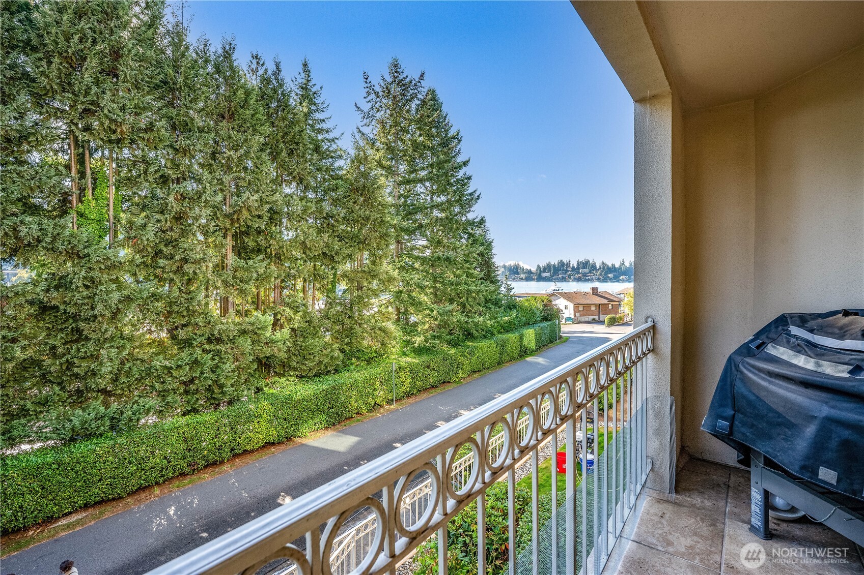 Condo balcony overlooking evergreen trees, nearby homes, and water in the distance.