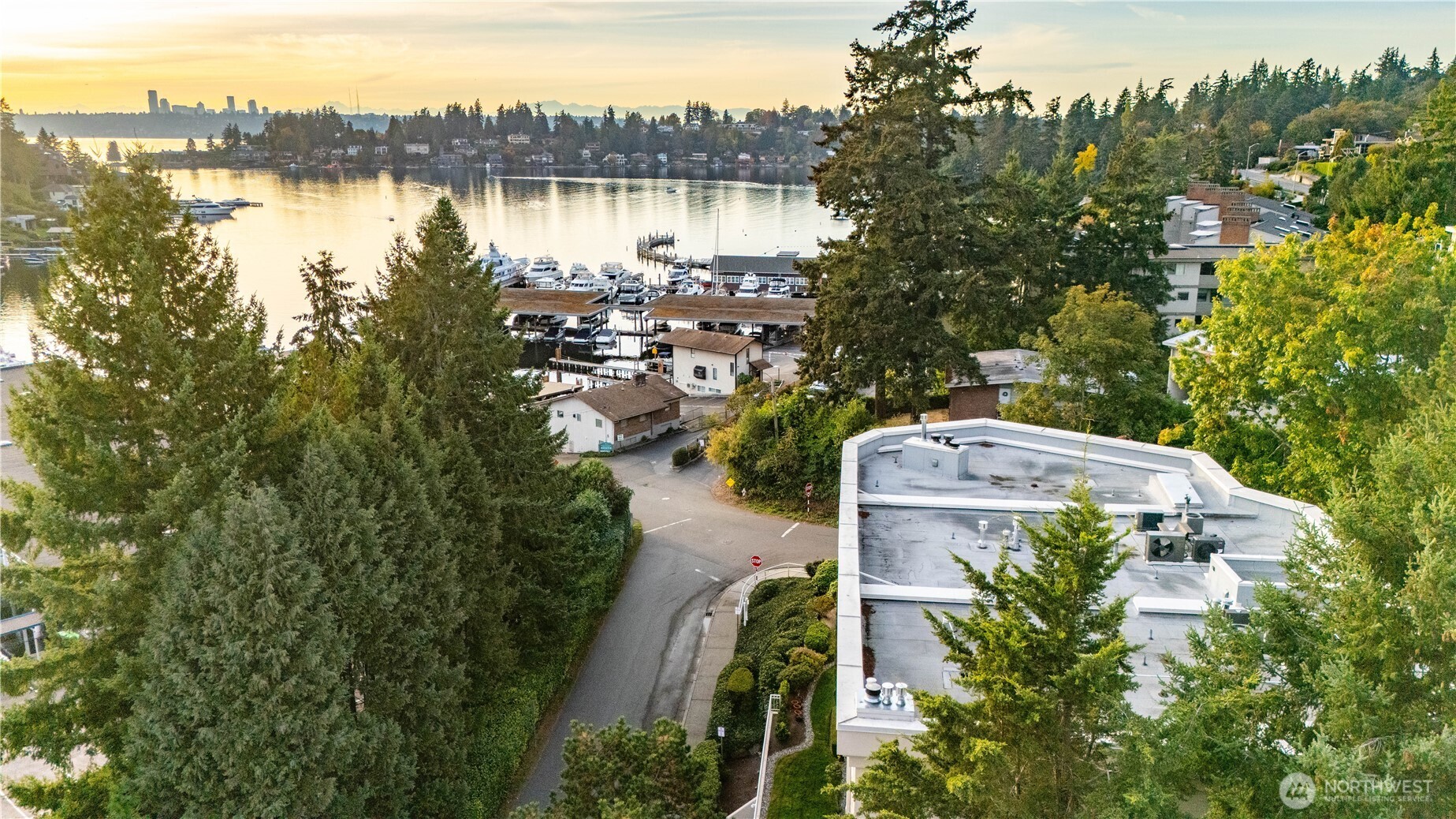 Aerial view over a marina and wooded shoreline toward a distant city skyline at sunset.