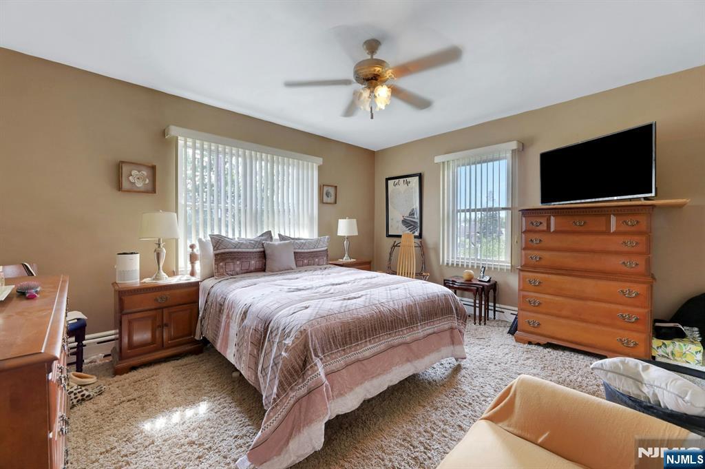 Furnished bedroom with a large bed, wooden dresser, wall-mounted television, and two bright windows.
