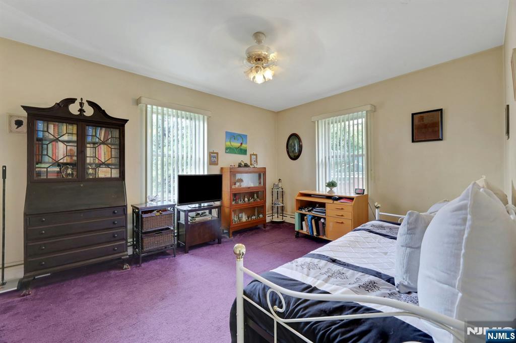 Bedroom with purple carpet, white metal bed, dark glass-front cabinet, and two windows with vertical blinds.