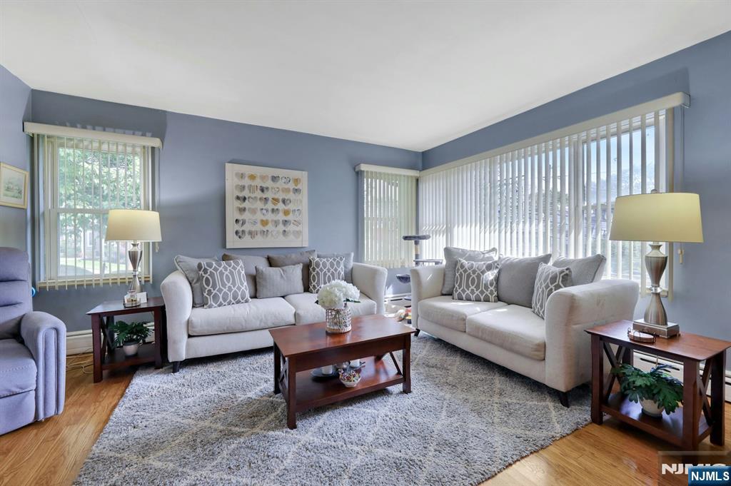 Living room with white sofas, a lavender armchair, and tall corner windows with vertical blinds.