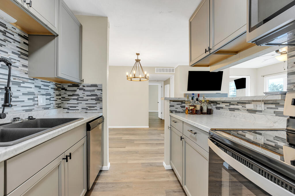 Renovated kitchen with gray cabinets, mosaic backsplash, stone counters, and a breakfast bar opening to the living area.
