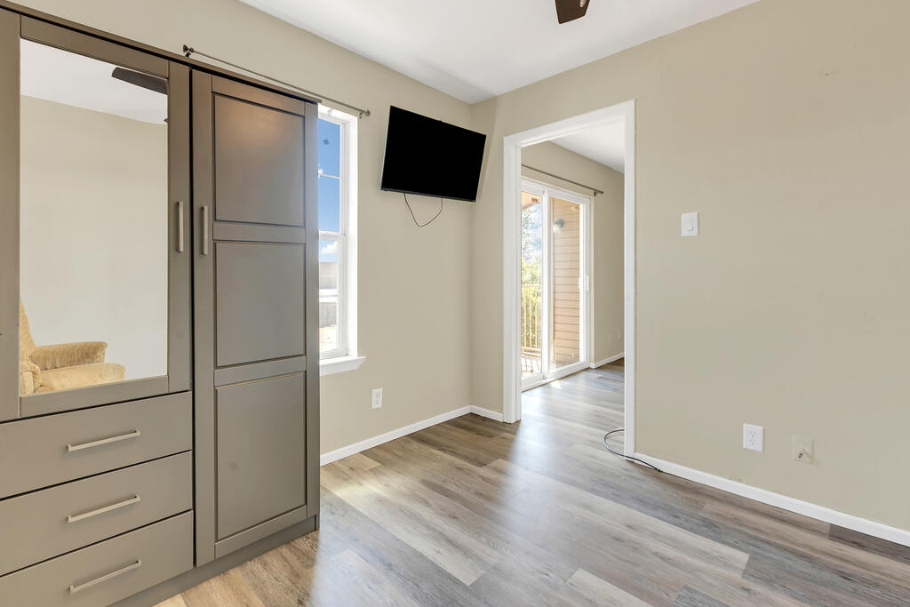 Mostly empty bedroom with a mirrored wardrobe, wall-mounted TV, and doorway to a sunlit adjoining room.