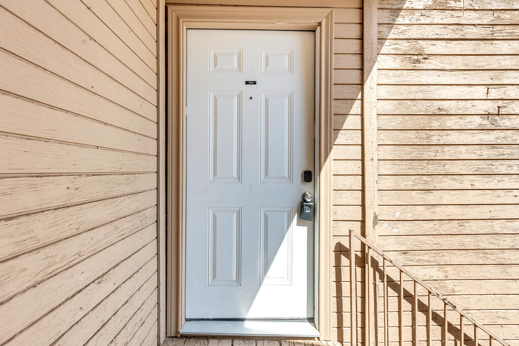 White paneled entrance door marked 704, set in tan siding beside a metal stair railing.