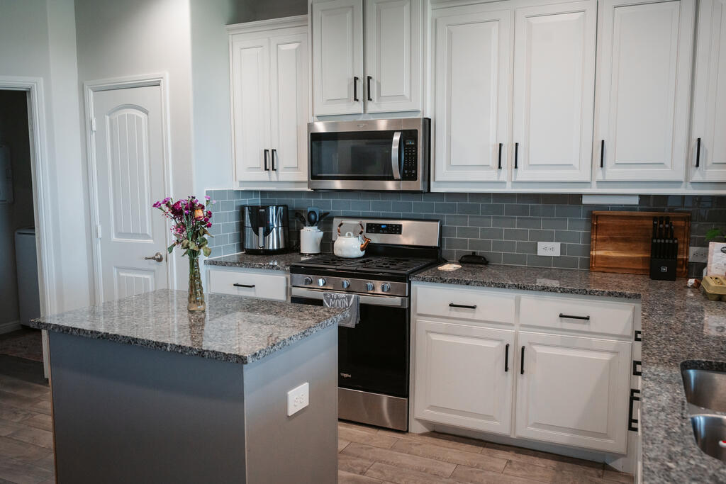 Kitchen with white cabinets, gray tile backsplash, stainless steel range, and a large granite-topped island.