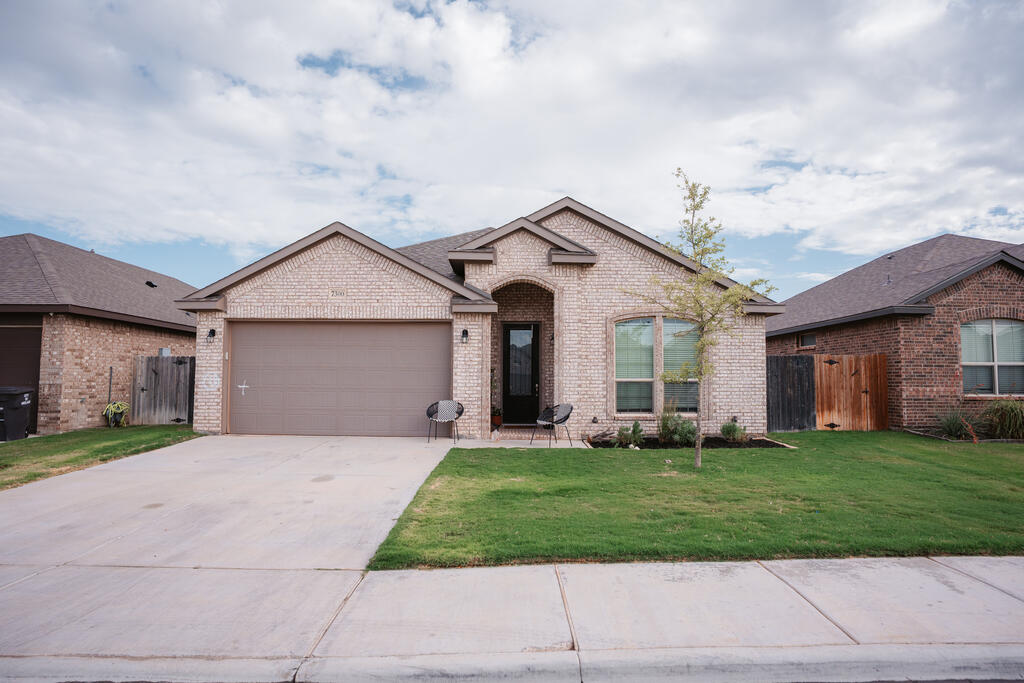 Light brick single-story house with a two-car garage, covered entry, and young tree in the front lawn.
