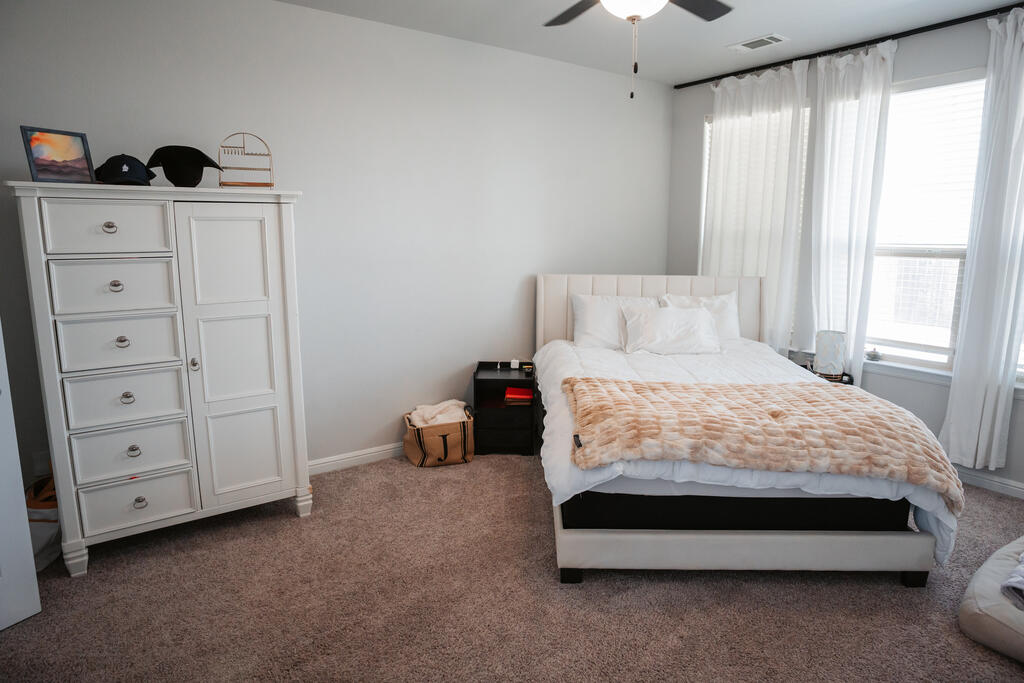 Furnished bedroom with a white upholstered bed, peach throw, tall curtained windows, and white dresser.