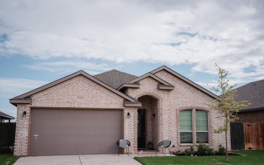 Brick single-story house with a two-car garage, arched entry, and two chairs by the front door.