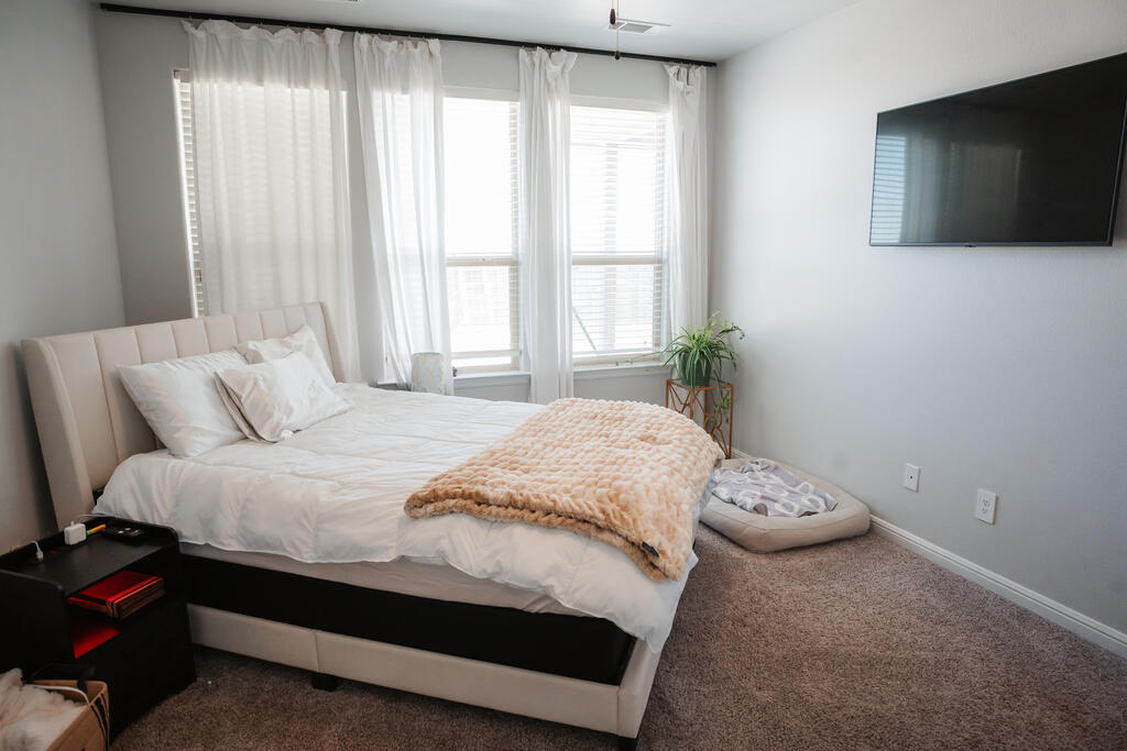 Bright bedroom with a large bed, curtained windows, wall-mounted TV, and carpet.