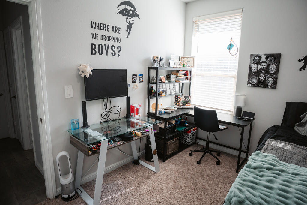 Bedroom with a gaming desk setup, shelving, and a bed beside a bright window.
