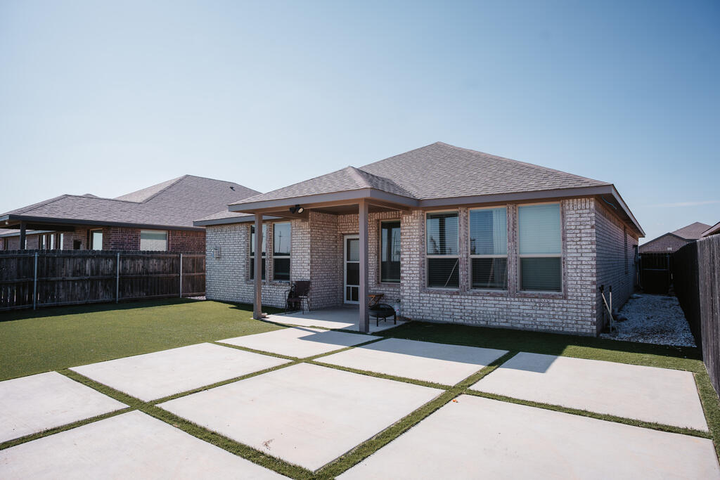 Brick home’s backyard with a covered patio, concrete squares, and strips of artificial turf.