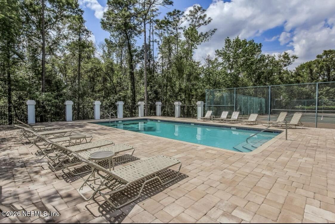 Outdoor pool and fenced tennis court surrounded by trees, with lounge chairs on the paved deck.