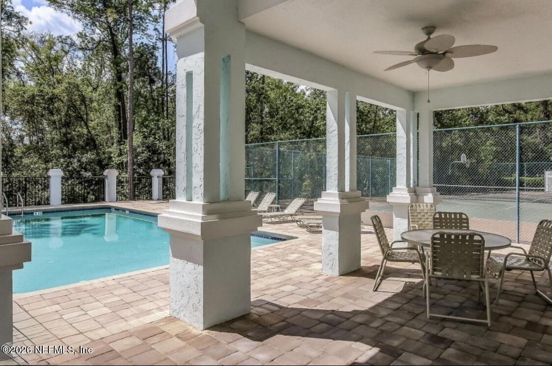 Community pool beside a covered patio with outdoor seating and a fenced tennis court.