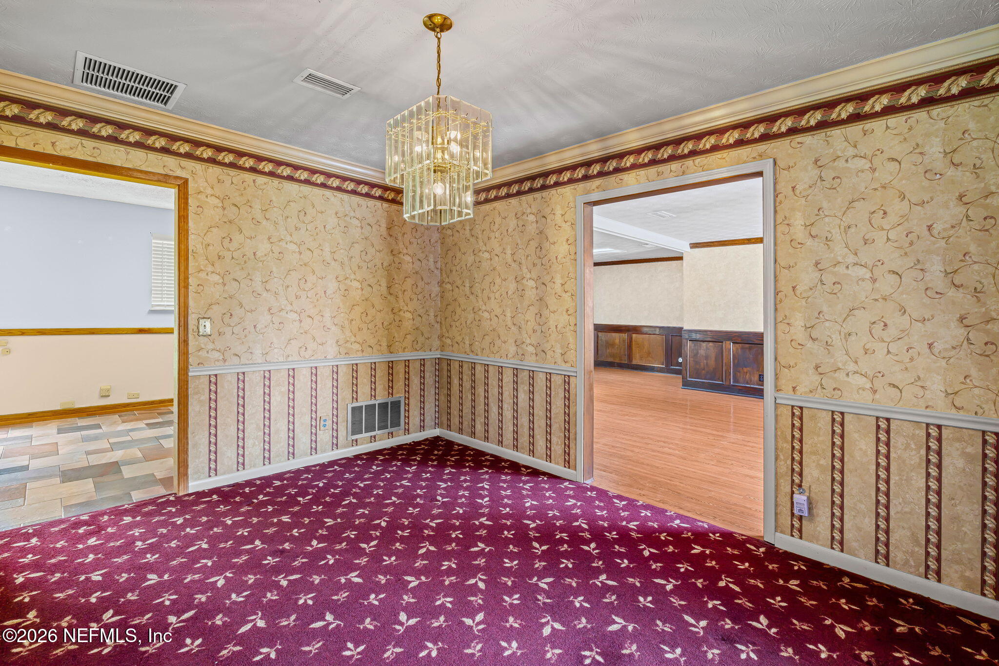 Empty formal dining room with ornate wallpaper, burgundy patterned carpet, and a crystal chandelier.
