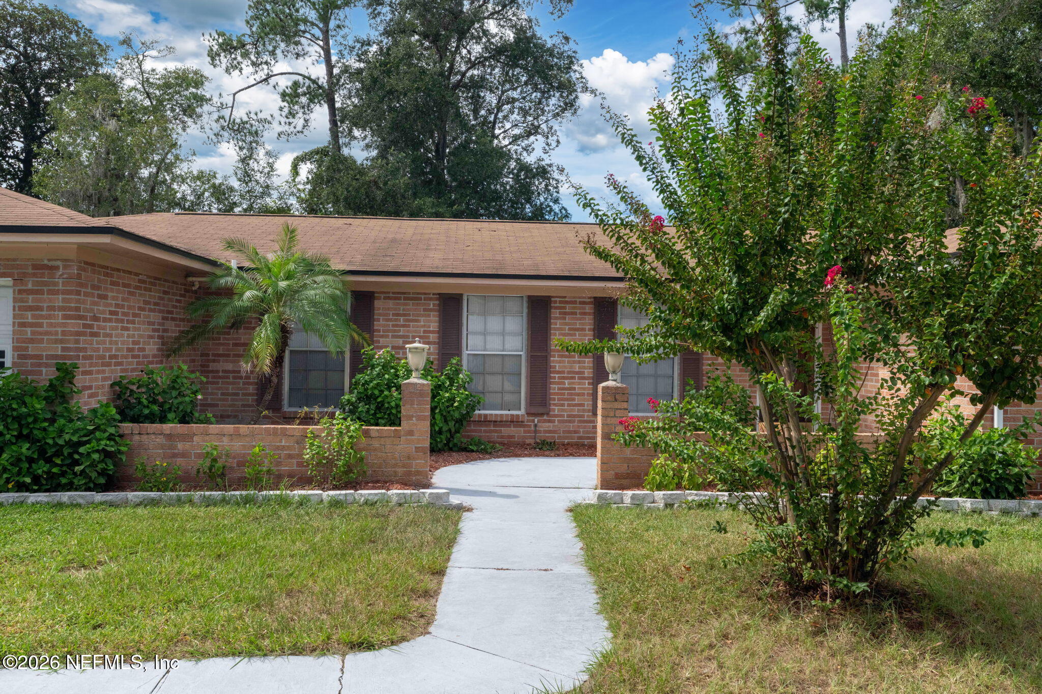 Single-story brick home with a concrete front walkway, low brick pillars, and flowering shrubs.