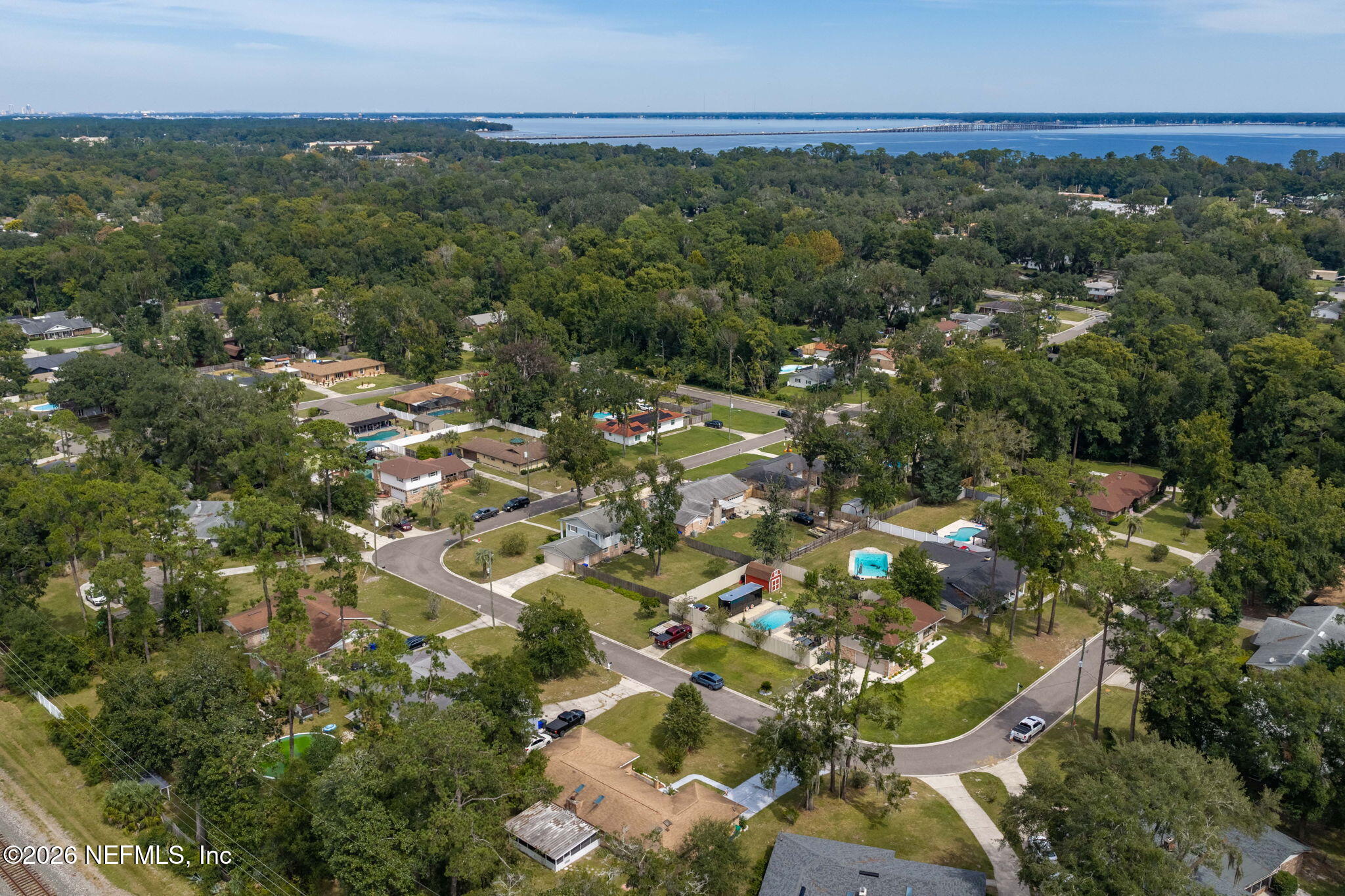 Aerial neighborhood view with tree-lined streets and homes, with water and a bridge in the distance.