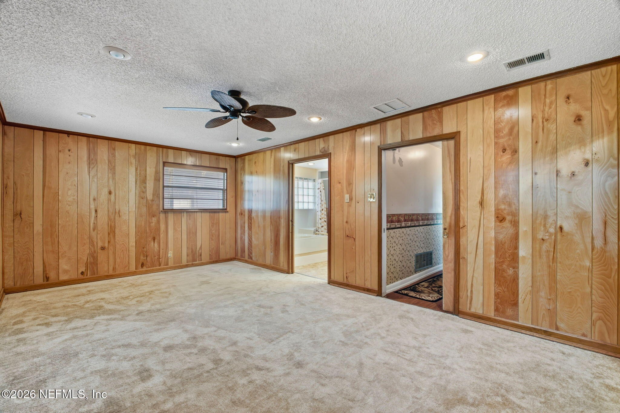 Empty bedroom with wood-paneled walls, beige carpet, ceiling fan, and doorways to adjoining rooms.
