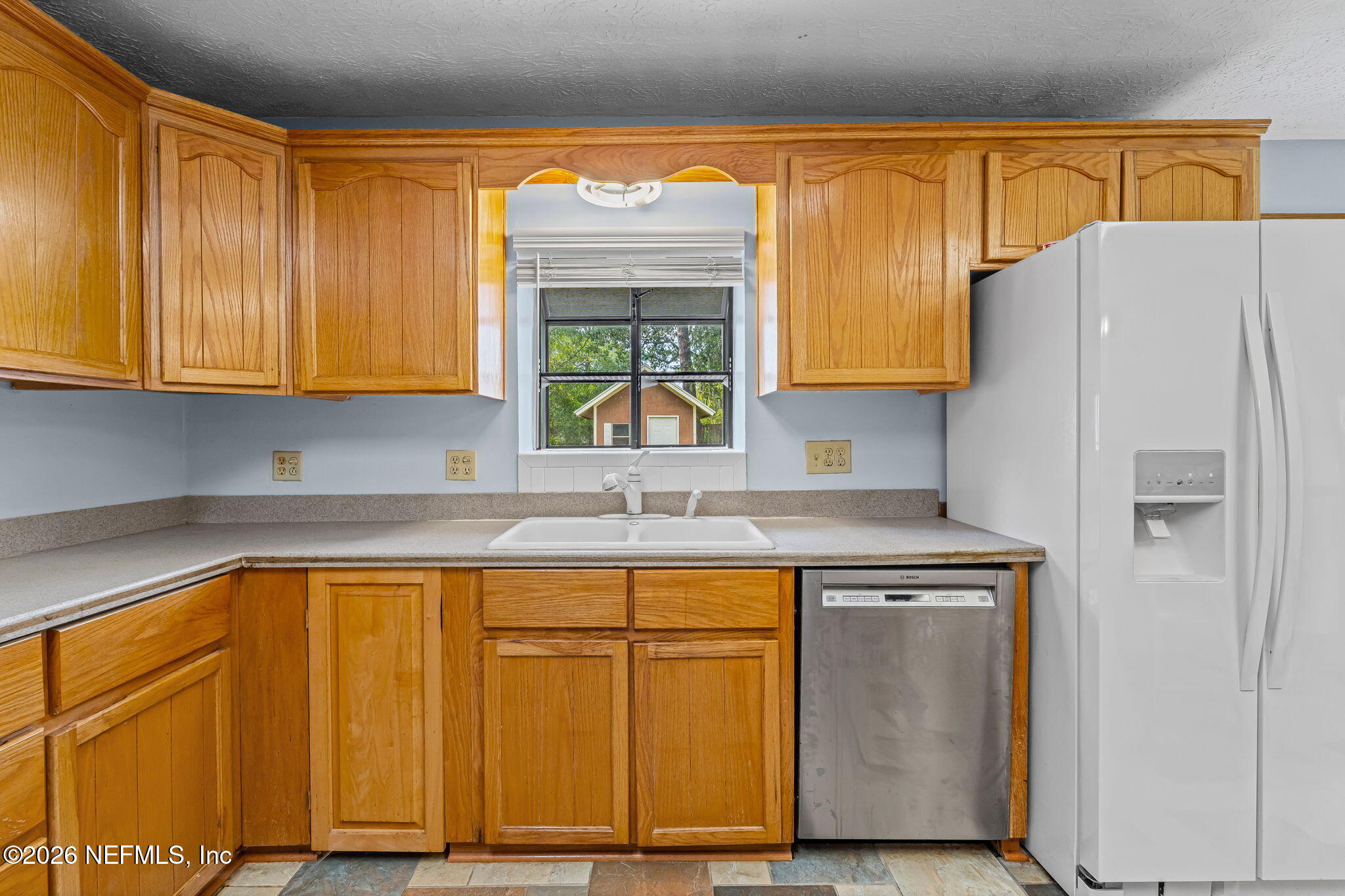 Kitchen with honey-colored cabinets, a double sink beneath a window, stainless-steel dishwasher, and white refrigerator.