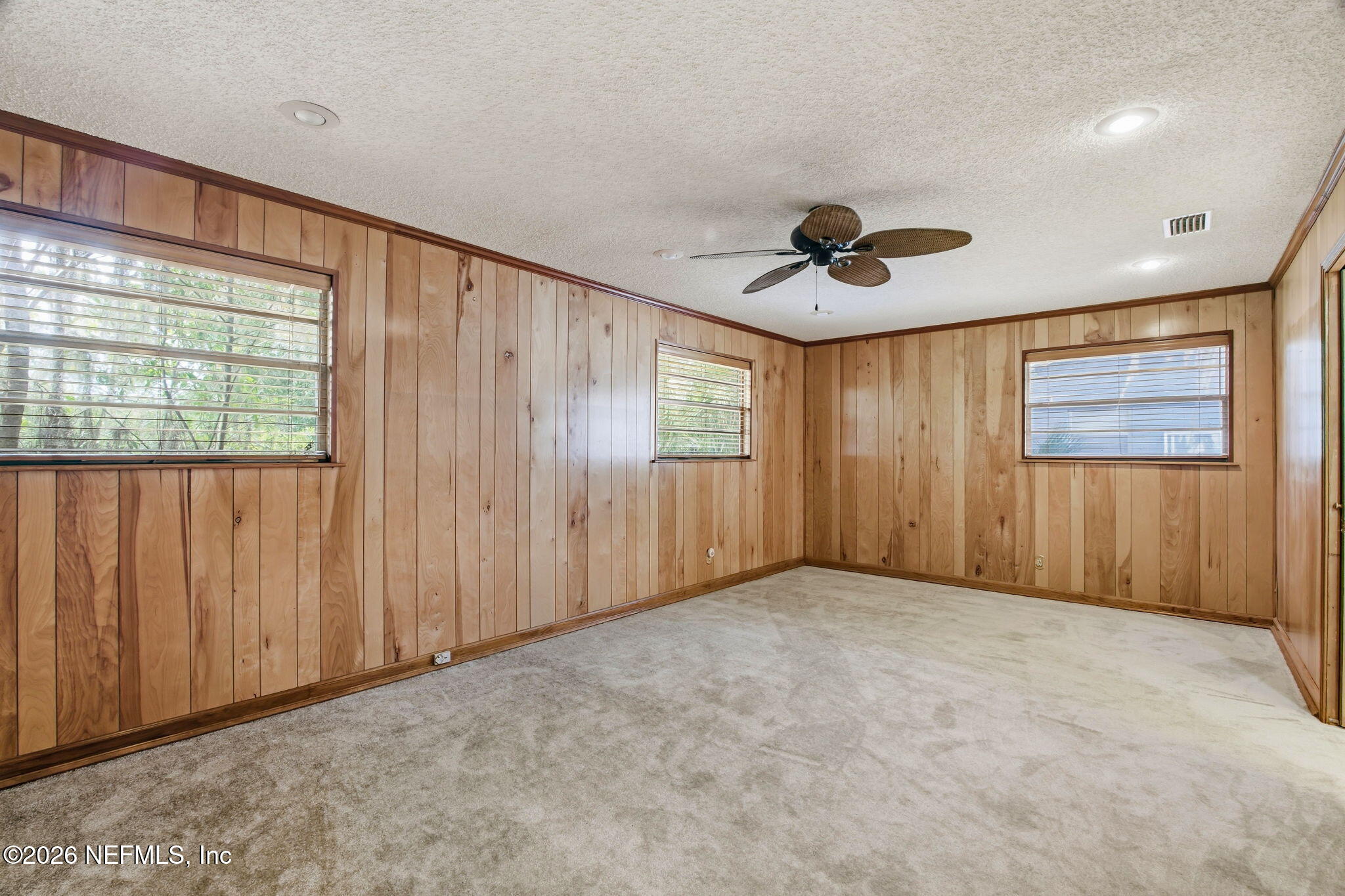 Vacant room with wood-paneled walls, carpet, three windows, and a ceiling fan.