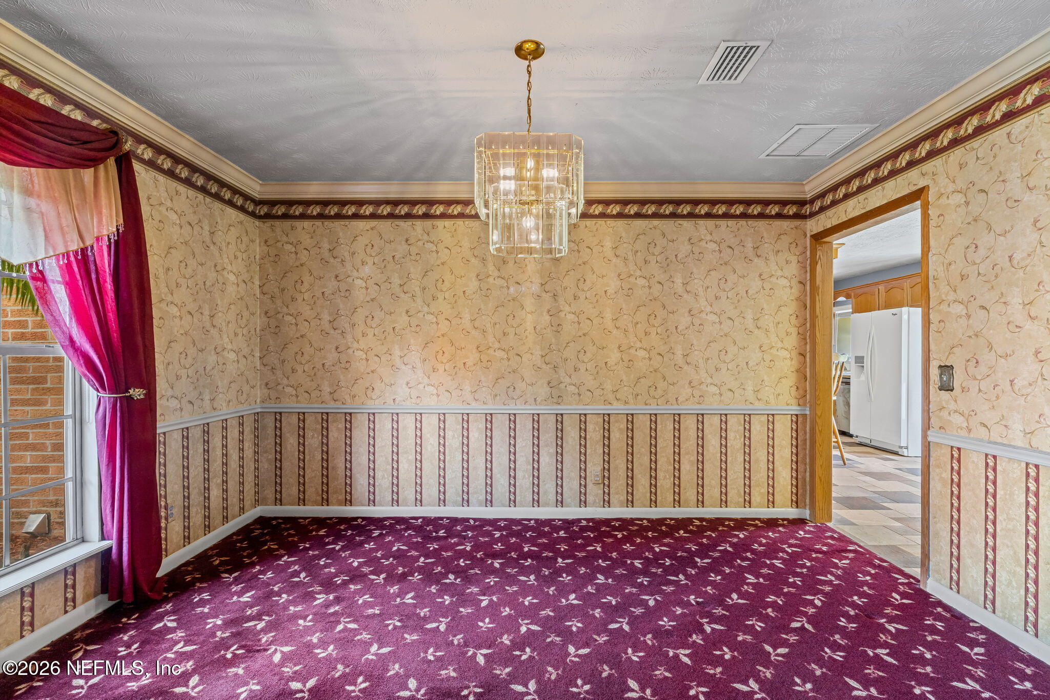 Empty dining room with burgundy patterned carpet, ornate wallpaper, and a crystal chandelier.