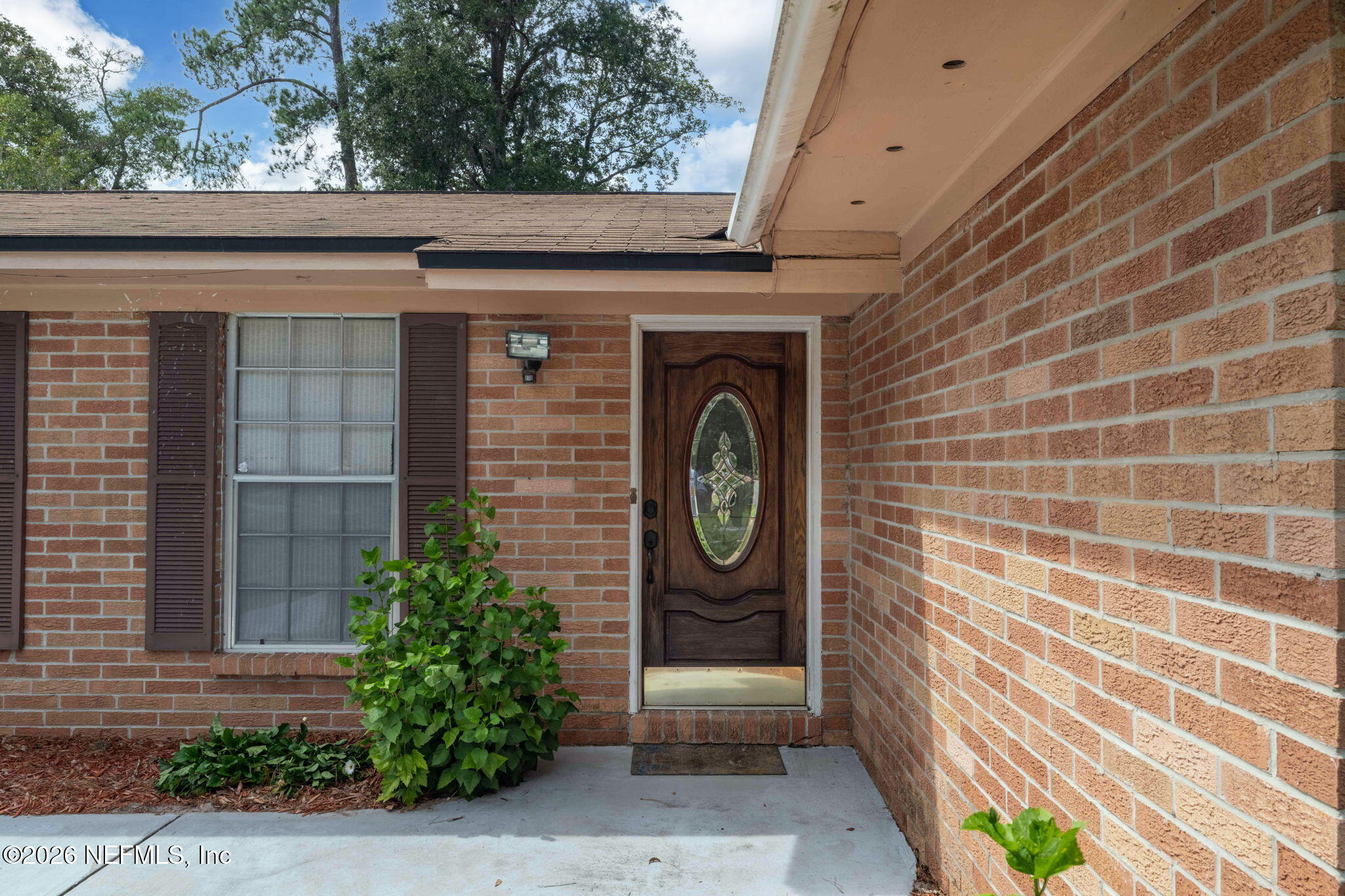 Brick home’s covered entry with an oval glass front door, shuttered window, and leafy plants.