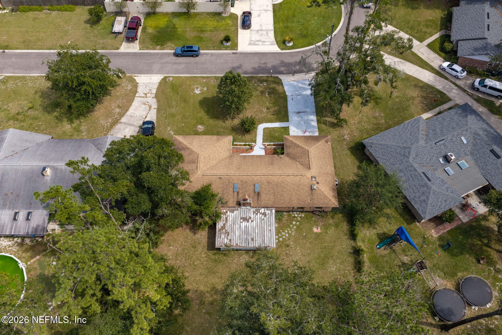 Aerial view of a tan-roofed home with a large backyard, covered patio, playset, and trampolines.