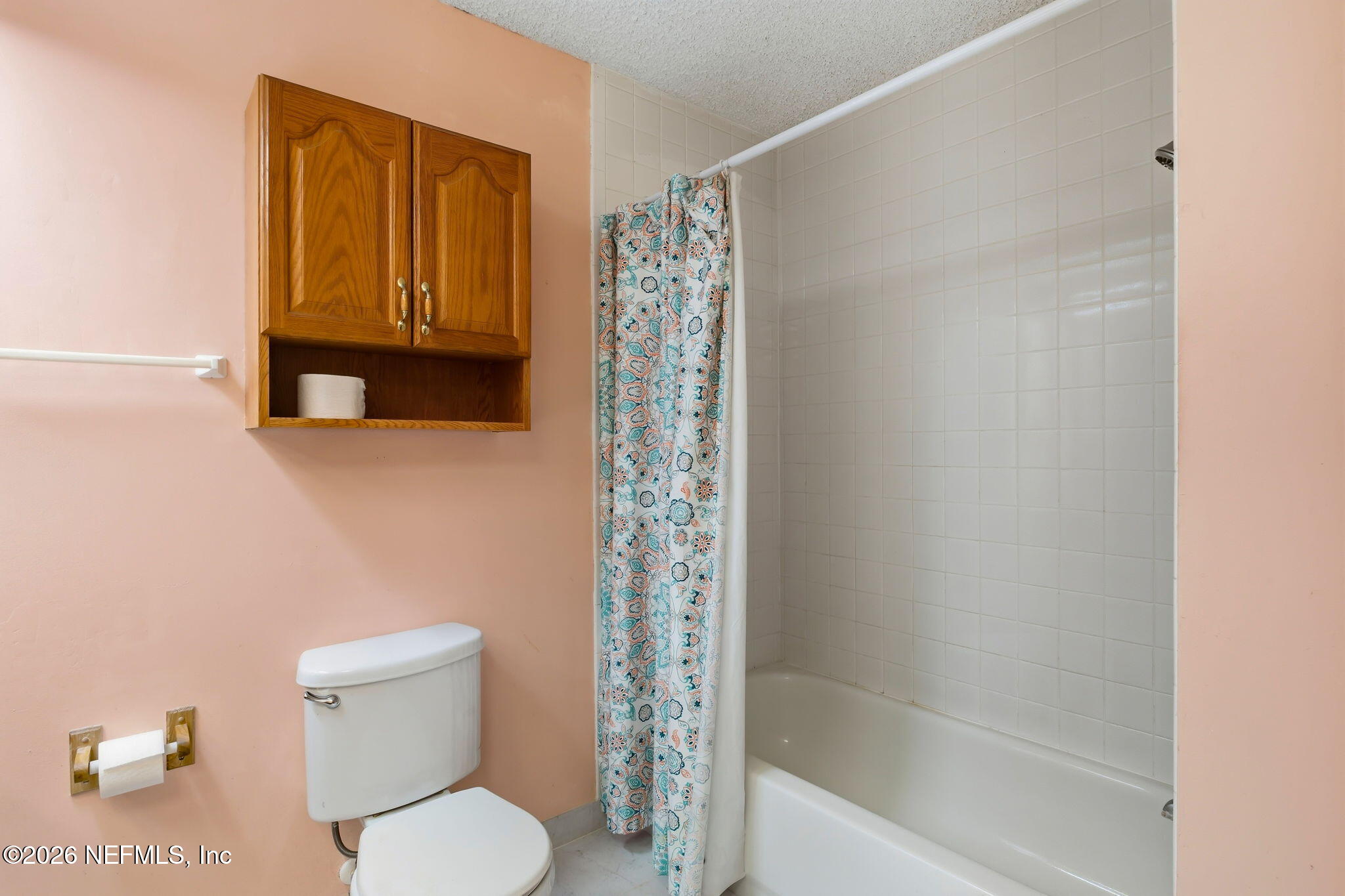 Bathroom with a tiled tub and shower, patterned curtain, white toilet, and wooden wall cabinet.
